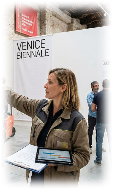 From Concept to Implementation A female Cultural Project Manager in a tan utility jacket holding a clipboard and a tablet with a project schedule. She is pointing toward an installation process in a large museum hall. In the background, a large banner reads 'VENICE BIENNALE' and a sign titled 'LOGISTICS & CUSTOMS' is visible near a group of technicians, illustrating the complex organization of an international art exhibition.
